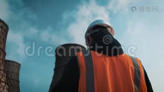 专业工程师戴着白色头盔和夹克看着工厂的管道。 冷却塔上有白色的视频