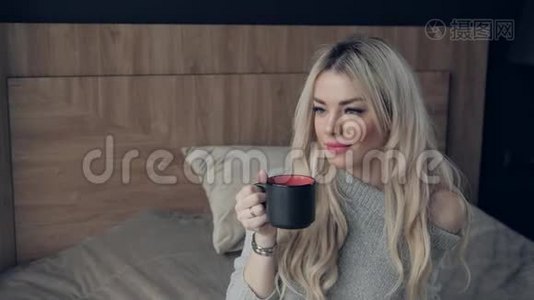 美丽的金发女人拿着陶瓷杯的茶或咖啡享受。 在床上喝茶。 看着窗户喝茶视频