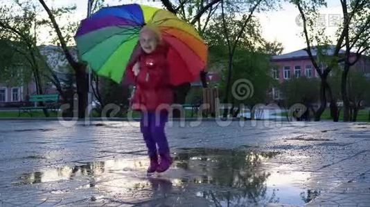 可爱的小女孩带着五颜六色的雨伞在日落时在公园的水坑里跳。视频