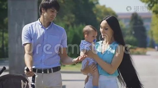 年轻的一家人抱着一个小男孩走在城市公园里。视频