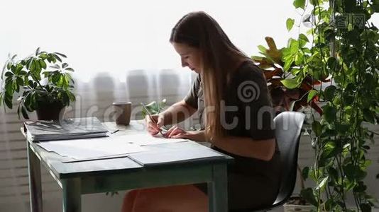 在家工作。 年轻漂亮的自由职业者正在房子窗户旁的桌子旁工作。 现代生态视频