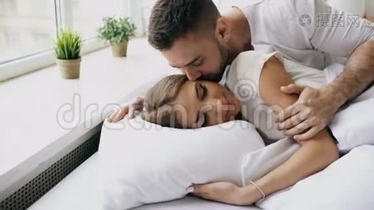 年轻美丽可爱的一对夫妇早上醒来。 迷人的男人在床上亲吻拥抱妻子视频