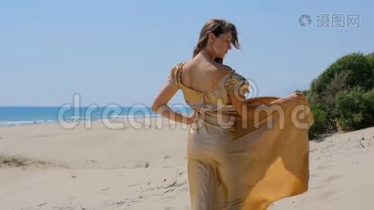 年轻漂亮的女人穿着金色的长裙站在靠近大海的沙丘上。视频