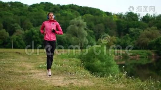 一个穿着粉红色夹克和黑色裤子的女孩戴着耳机在河边跑步，为马拉松做准备视频