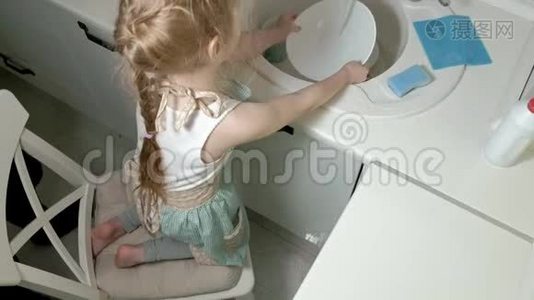 穿着围裙的漂亮小女孩站在椅子上，在明亮的厨房里，用海绵洗碗，帮助视频