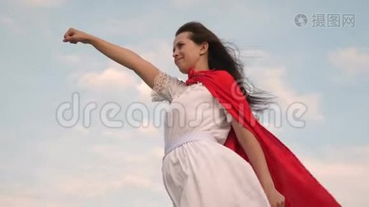 美丽的超级英雄女孩站在一片红色斗篷的田野上，斗篷在风中飘扬。 女孩梦想成为一个视频