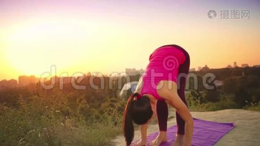 一个年轻的女人在大城市的背景下在山上练习瑜伽。 健康的女人在日落时做运动。 A.视频