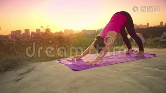 一个年轻的女人在大城市的背景下在山上练习瑜伽。 健康的女人在日落时做运动。 A.视频