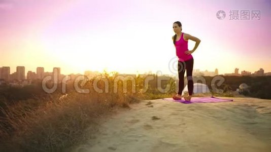 一个年轻的女人在大城市的背景下在山上练习瑜伽。 健康的女人在日落时做运动。 A.视频