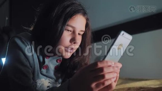 十几岁的女孩晚上在床上看她的智能手机。 一个小女孩在社交中写了一条聊天信息视频