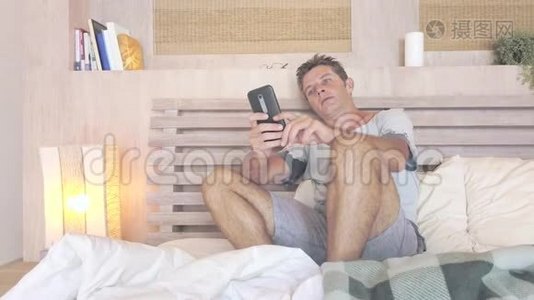 年轻有魅力的30岁男人，蓝眼睛，在家里，躺在床上，用手机上的网络社交媒体约会应用程序快乐放松视频