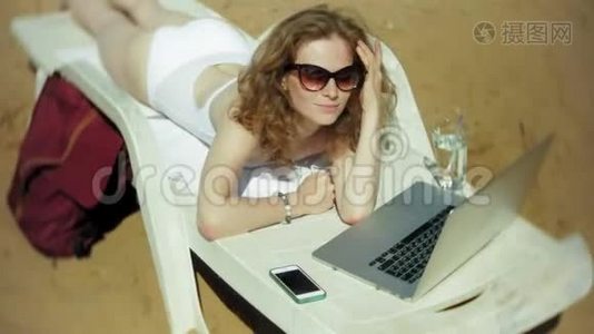 一个穿着白色比基尼的年轻女孩躺在沙滩上的躺椅上晒太阳，正在使用笔记本电脑视频