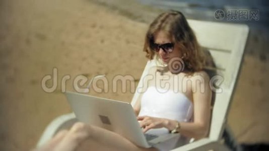一个穿着白色比基尼的年轻女孩躺在沙滩上的躺椅上晒太阳，正在使用笔记本电脑视频