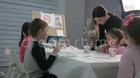 孩子们，男孩和女孩在教室里围着桌子坐在一起画画。 与他们在一起的是他们的年轻视频
