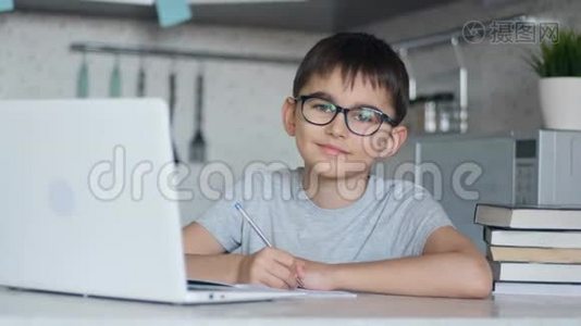 戴眼镜的孩子在家里坐在餐桌旁的厨房里，用笔记本电脑、书和笔记本上课视频