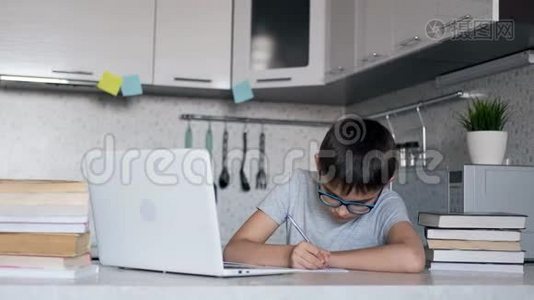 戴眼镜的孩子在家里坐在餐桌旁的厨房里，用笔记本电脑、书籍和笔记本上课。视频