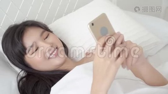 美丽的亚洲年轻女性，看着摸着智能手机，在卧室的床上享受社交网络视频