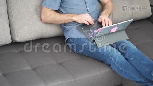 自信的年轻人坐在家里的沙发上，打开并开始使用笔记本电脑。 家庭在线工作委员会。 病毒检疫视频