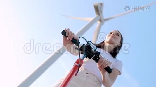 女孩带着电动滑板车在一个巨大的旋转涡轮的风力发电机的背景。视频