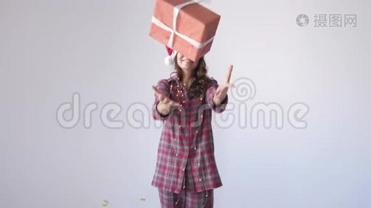 穿着圣诞帽和睡衣的快乐女孩在白色背景下扔礼物盒等待假期。 慢动作视频