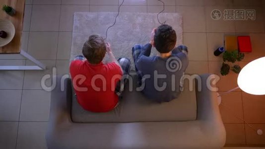 两个穿睡衣的年轻人用操纵杆玩电子游戏，在客厅里互相交谈。视频