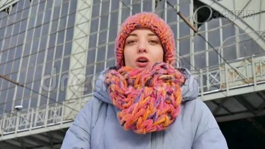一个年轻漂亮的成年女孩，穿着蓝色外套，戴着五颜六色的帽子和围巾，在阳光明媚的火车站的冬天肖像。视频