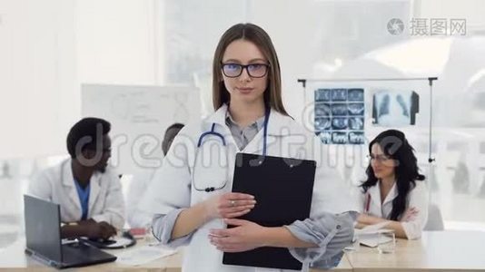 年轻的女医生穿着白色外套，听诊器微笑着，在诊所的同事背景上看着相机视频