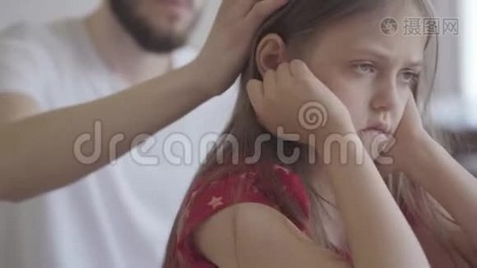 特写一幅悲伤的小女孩坐在前景上的肖像，双手紧紧地抱着她的头。 他的父亲视频