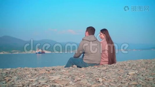 一个年轻的女孩和一个男人在春天的海滩上坐着说话。 那个女孩和那个相框里的家伙旋转。视频