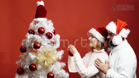 戴红帽子的长胡子男人拥抱一个装饰圣诞树的女人。 可爱的夫妇装饰圣诞树视频