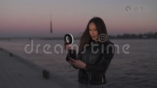 一位年轻的女士在日落时分用一个环形闪光灯作为补光灯自拍，可以看到多加瓦河视频