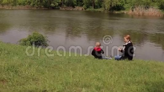 妈妈和儿子正坐在春天河岸上的绿草上。视频