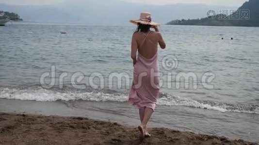 穿着粉红色丝绸裙子和草帽的女人在海滩上散步视频
