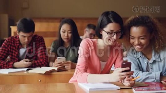 漂亮的女学生正在使用智能手机，看屏幕，有说有笑，坐在大学的桌子上。 社会视频