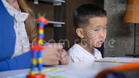 亚洲小男孩从地板上拿着落下来的笔，和母亲一起学习，坐在桌子、书架和砖墙后面视频