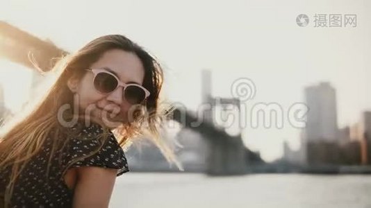 快乐的欧洲女孩戴着太阳镜微笑，在纽约布鲁克林大桥享受令人惊叹的河流日落，看着相机。视频
