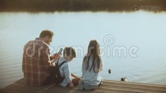 父亲和两个孩子坐在湖边码头上。 欧洲男人带着孩子用智能手机拍摄水鸟。 4K.视频