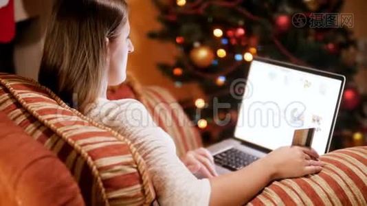4k镜头显示，年轻女子坐在扶手椅旁边的发光圣诞树使用笔记本电脑进行网上购物。 购买视频