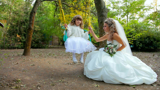 在公园里穿着婚纱的新娘和一个坐在秋千上的小女孩视频