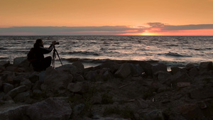 在日落时分摄影师站在岩石上用三脚架上的相机拍摄海边的全景照片24秒视频