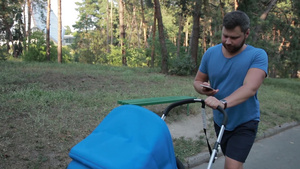 年轻的嬉皮士父亲穿着蓝色T恤和短裤，忙于智能手机在公共公园散步，推着婴儿车和蹒跚学步的儿子。 时尚的爸爸，留着18秒视频