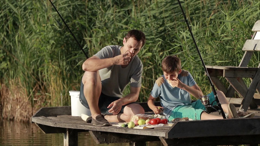 父亲和儿子在池塘边的木墩上吃早餐[小木凳]视频