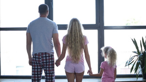 积极的家庭与可爱的卷发金发女儿穿着睡衣手拉手站在大窗户旁10秒视频