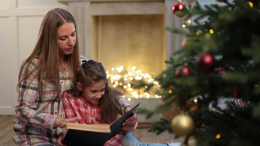 漂亮的年轻母亲在圣诞树附近给她可爱的女儿读书视频