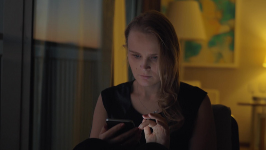 金发女人坐在室内玩智能手机[碧眼]视频