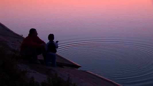 日落时分一队父子坐在河岸边向水里丢石头视频