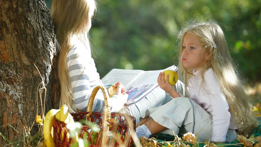 两个金发小女孩坐在树下在看书吃苹果享受阳光明媚的秋天视频
