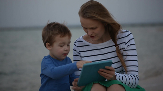 母亲和她的小儿子在海边玩平板电脑视频