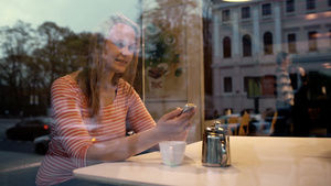 从咖啡馆外拍摄的年轻女人在咖啡馆里喝着咖啡玩着手机而玻璃里反射了城市生活16秒视频