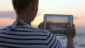 女士在日落时用平板电脑拍摄波涛汹涌的海浪的背景11秒视频